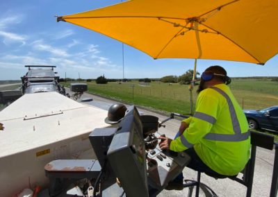 Road construction worker controlling a milling machine, shaded by a yellow umbrella in a rural area.