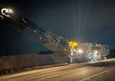 Asphalt milling machine working at night on a road construction project, illuminated by bright lights.