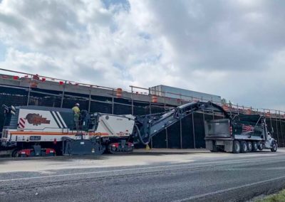 Construction crew using a milling machine to remove old asphalt near a newly constructed highway.