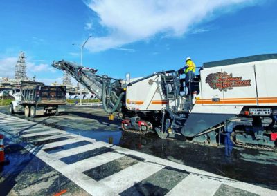 A road milling machine and dump truck during a road resurfacing project, with a crosswalk visible in the foreground.