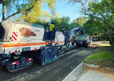 Milling machine operating on a tree-lined residential street with a dump truck and workers nearby.