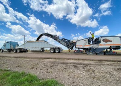 A milling machine operating on a rural road, loading debris into a dump truck.
