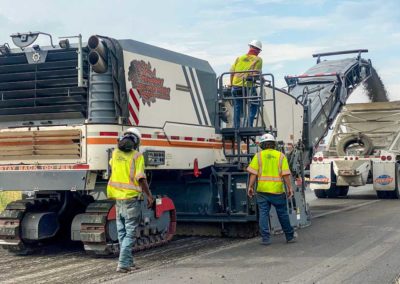 Three workers control a milling machine, loading milled asphalt into a truck for transport.