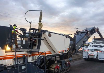 Evening road milling with workers and equipment actively removing the old road surface.
