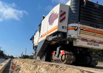 Milling machine working on a road edge, guided by workers, with a clear blue sky in the background.