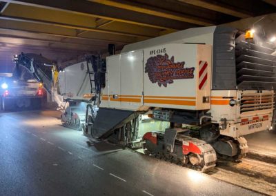 Nighttime milling operation under a low overpass with bright lights illuminating the site.