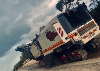 Road milling machine working on a sloped road under a cloudy sky.
